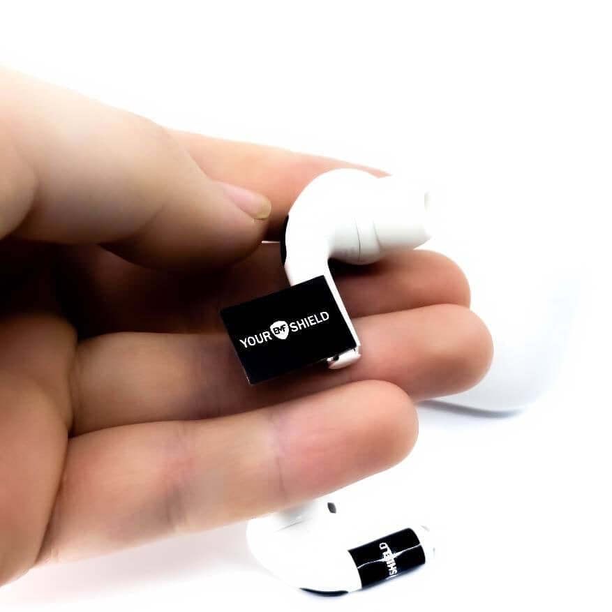 A hand holding a pair of white earbuds with an adhesive EMF shield attached to one of the earbuds.